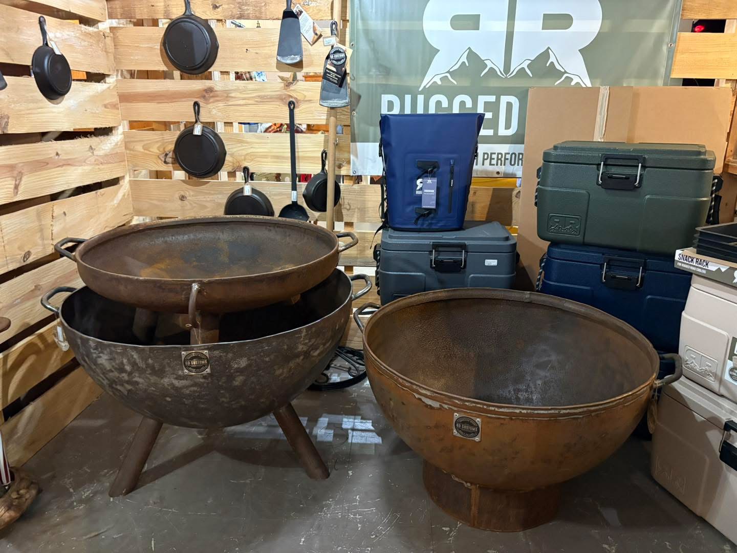 Large metal fire pits with coolers and cast iron cookware behind them.